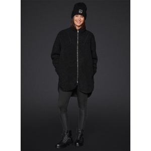A woman is stood facing forward, smiling while wearing the Mountain Horse Emma Pile Jacket in black, It is a mid thigh length jacket made from soft borg fleece to keep you warm and cosy during those winter yard days.